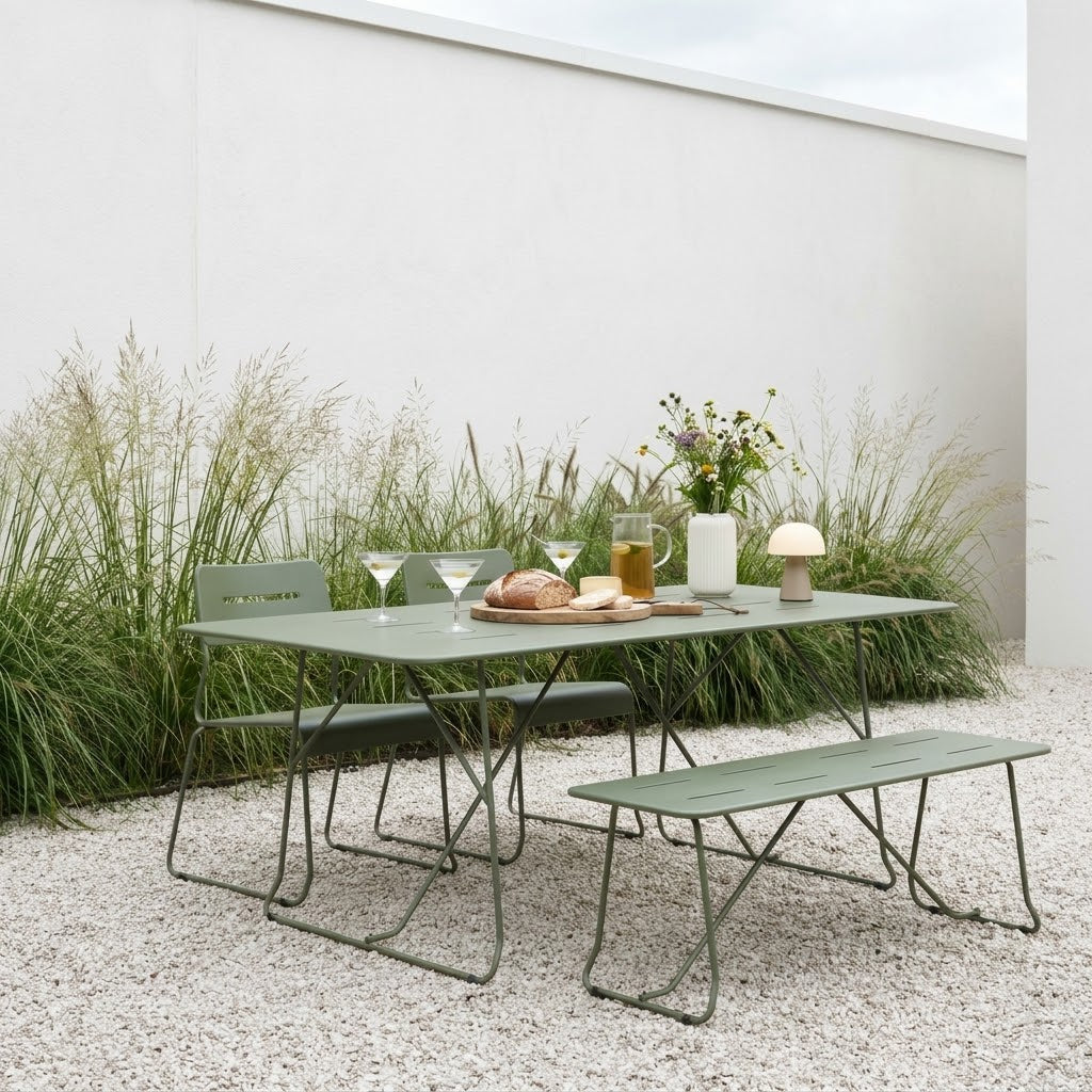 An outdoor dining set consisting of a rectangular metal table, a matching bench, and a chair, all in a muted sage green color. The set is styled on a gravel patio with tall ornamental grasses, a white wall, and dining tableware on the table.