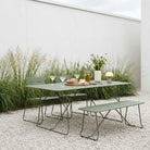 An outdoor dining set consisting of a rectangular metal table, a matching bench, and a chair, all in a muted sage green color. The set is styled on a gravel patio with tall ornamental grasses, a white wall, and dining tableware on the table.