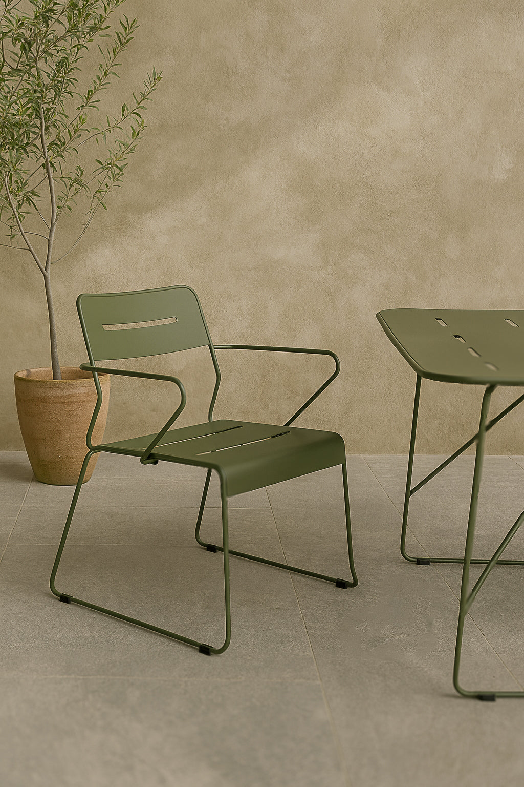 Olive green outdoor dining chair alongside a minimalist table, set against a textured beige wall with a potted plant in the background.